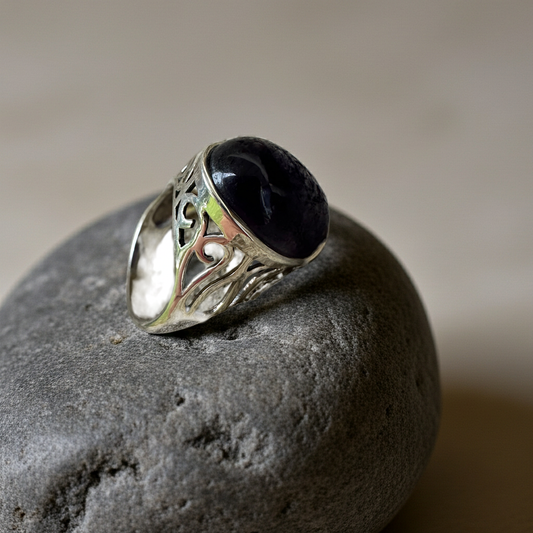 “Amethyst Whisper” Silver Ring with a dark polished amethyst stone and intricate silver filigree design resting on a smooth stone surface