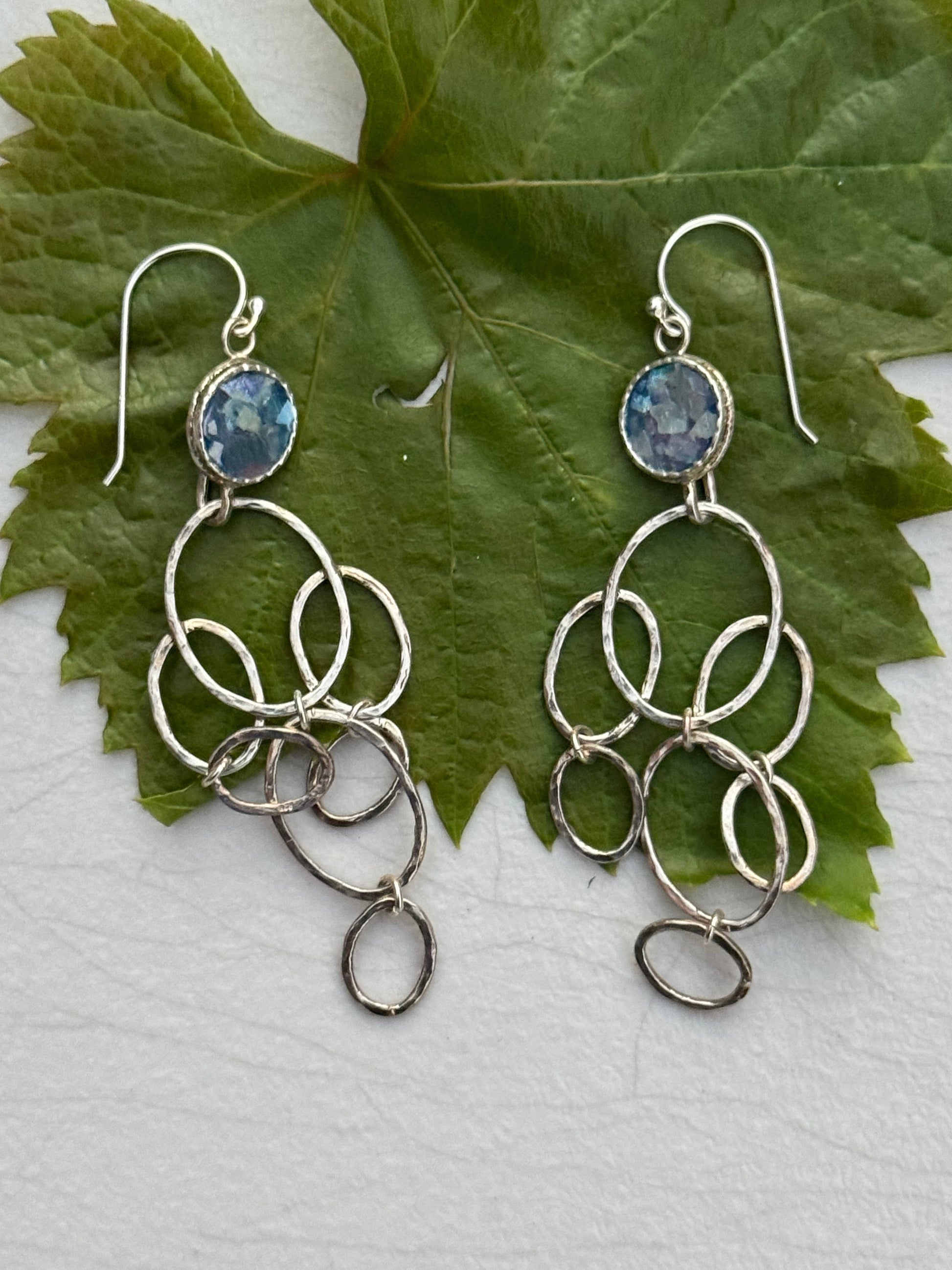 LONG ROMAN GLASS EARRINGS featuring silver interconnected loops with a blue glass stone at the top, displayed on a green leaf background.