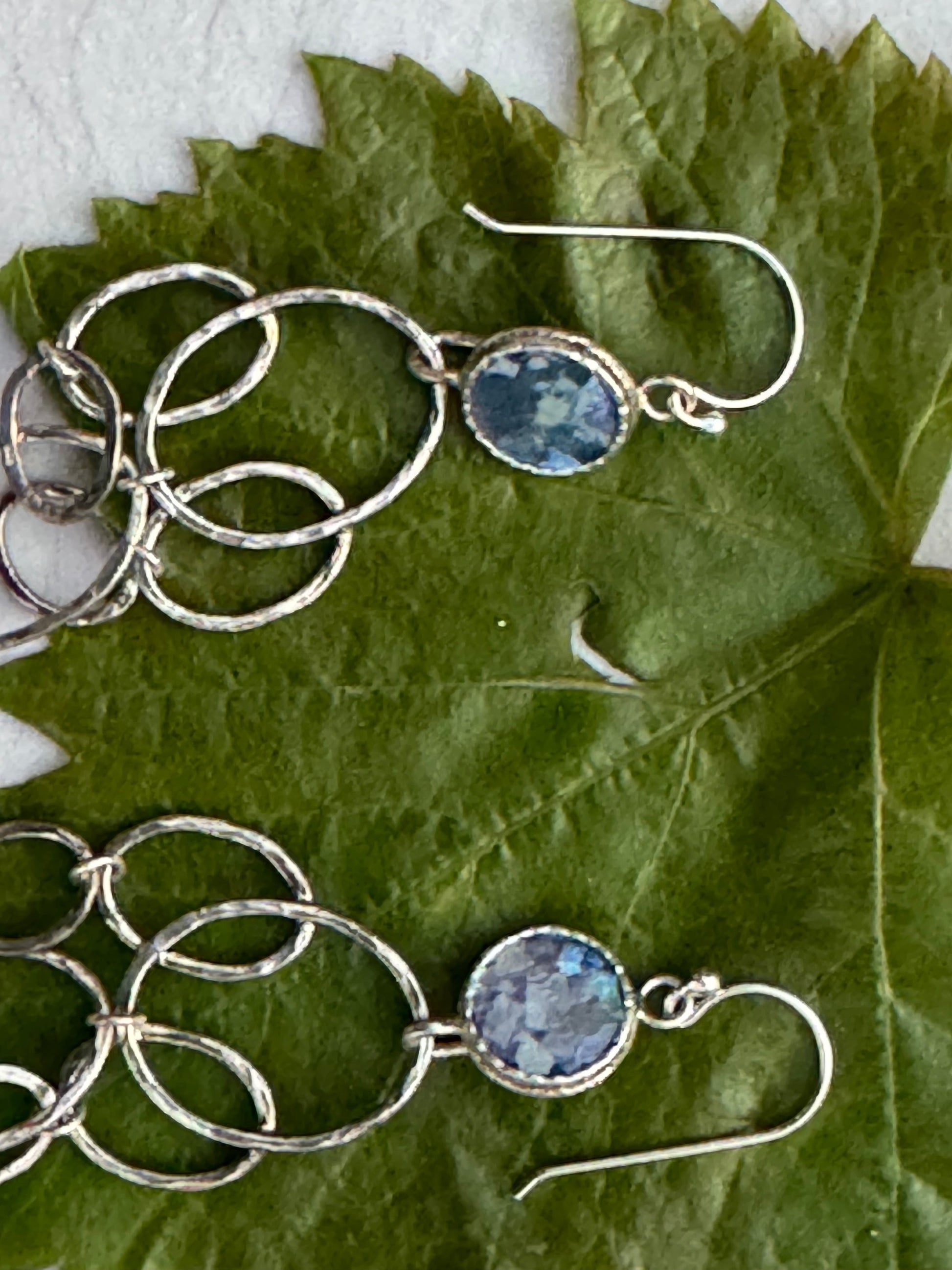 LONG ROMAN GLASS EARRINGS featuring linked hammered silver hoops with oval blue glass stones set in silver, displayed on a green leaf background.