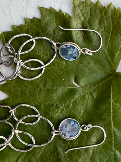 LONG ROMAN GLASS EARRINGS featuring linked hammered silver hoops with oval blue glass stones set in silver, displayed on a green leaf background.