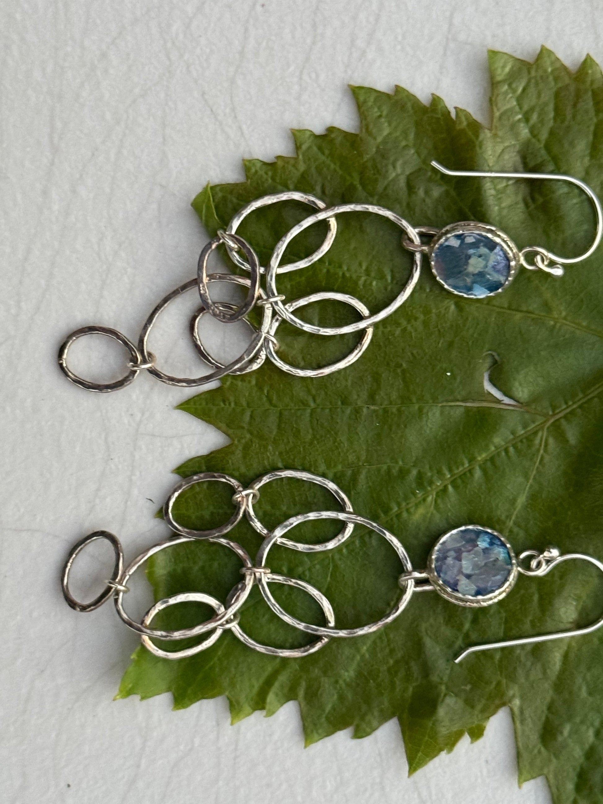 LONG ROMAN GLASS EARRINGS featuring silver interlocking oval loops with blue glass stones, displayed on a green leaf background, artisan craft jewelry