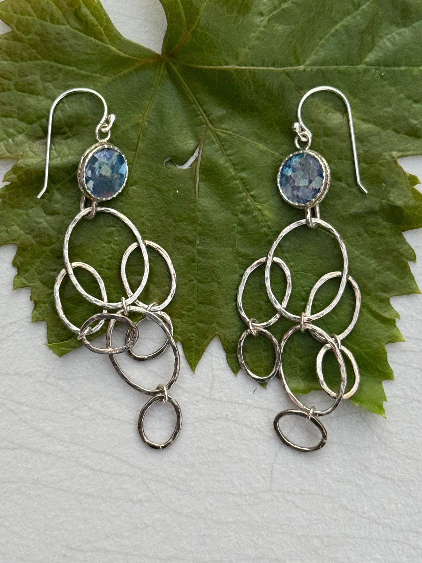 LONG ROMAN GLASS EARRINGS featuring silver loops and blue glass stones, displayed on a green leaf background, Mediterranean artisan design.