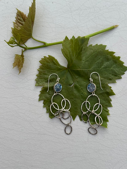 LONG ROMAN GLASS EARRINGS featuring multiple interlinked silver rings with blue glass stones, displayed on a large green leaf background.