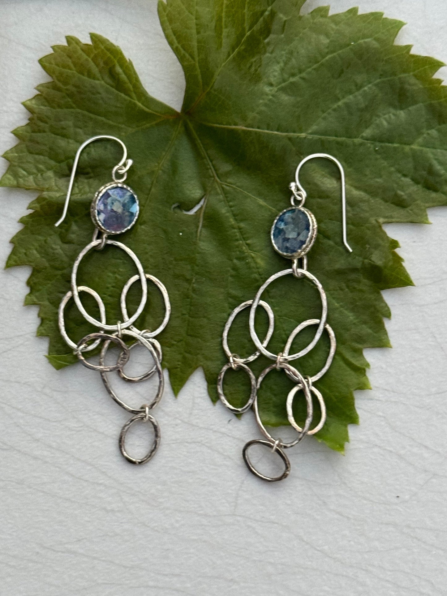 LONG ROMAN GLASS EARRINGS featuring silver hoops and a blue-toned glass stone, displayed on a large green leaf background, showcasing artisan craftsmanship.