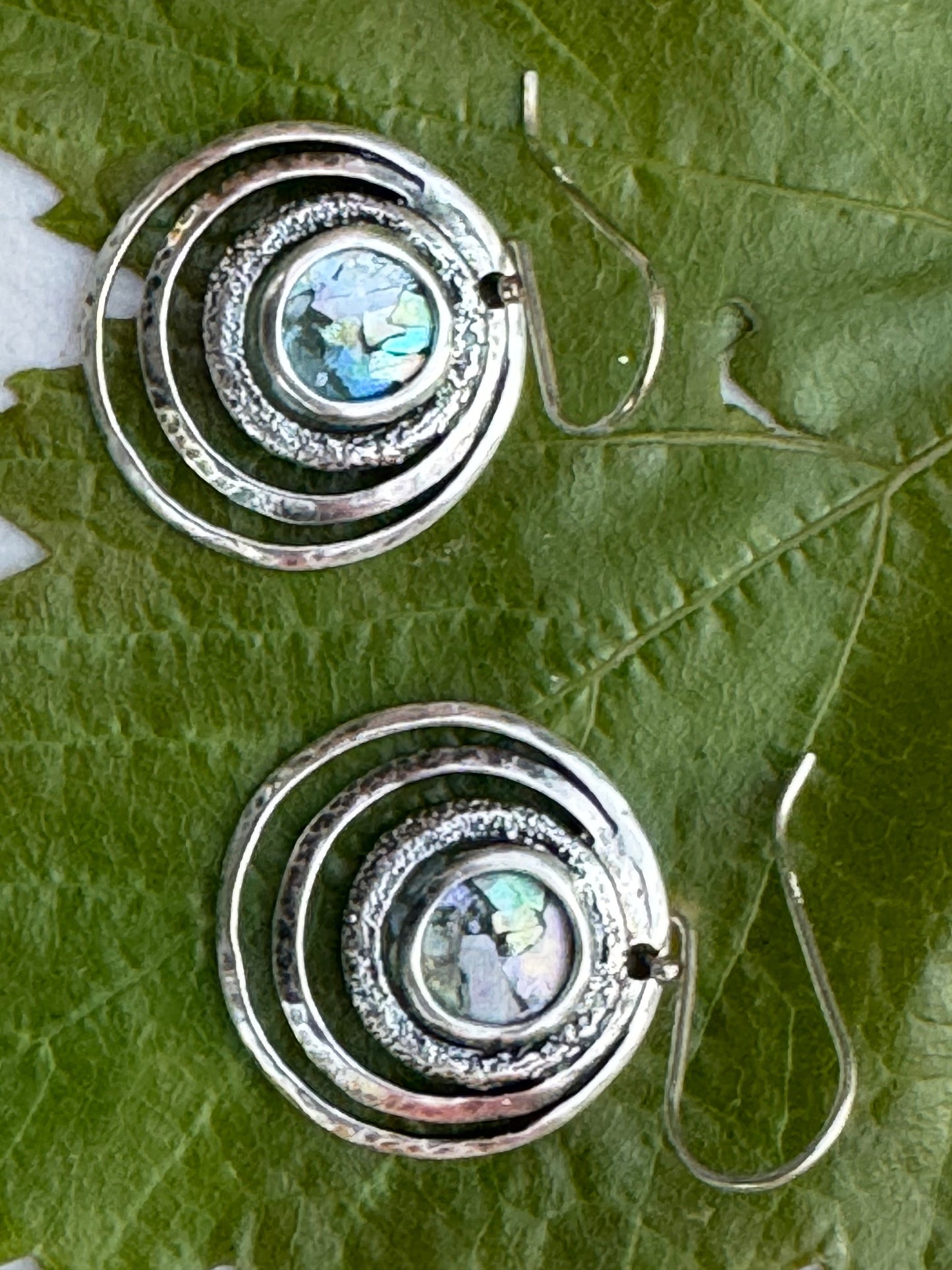 Round 3 Circles Ancient Roman Glass Sterling Earrings featuring concentric silver circles with textured detailing and a central iridescent ancient Roman glass piece on a green leaf background